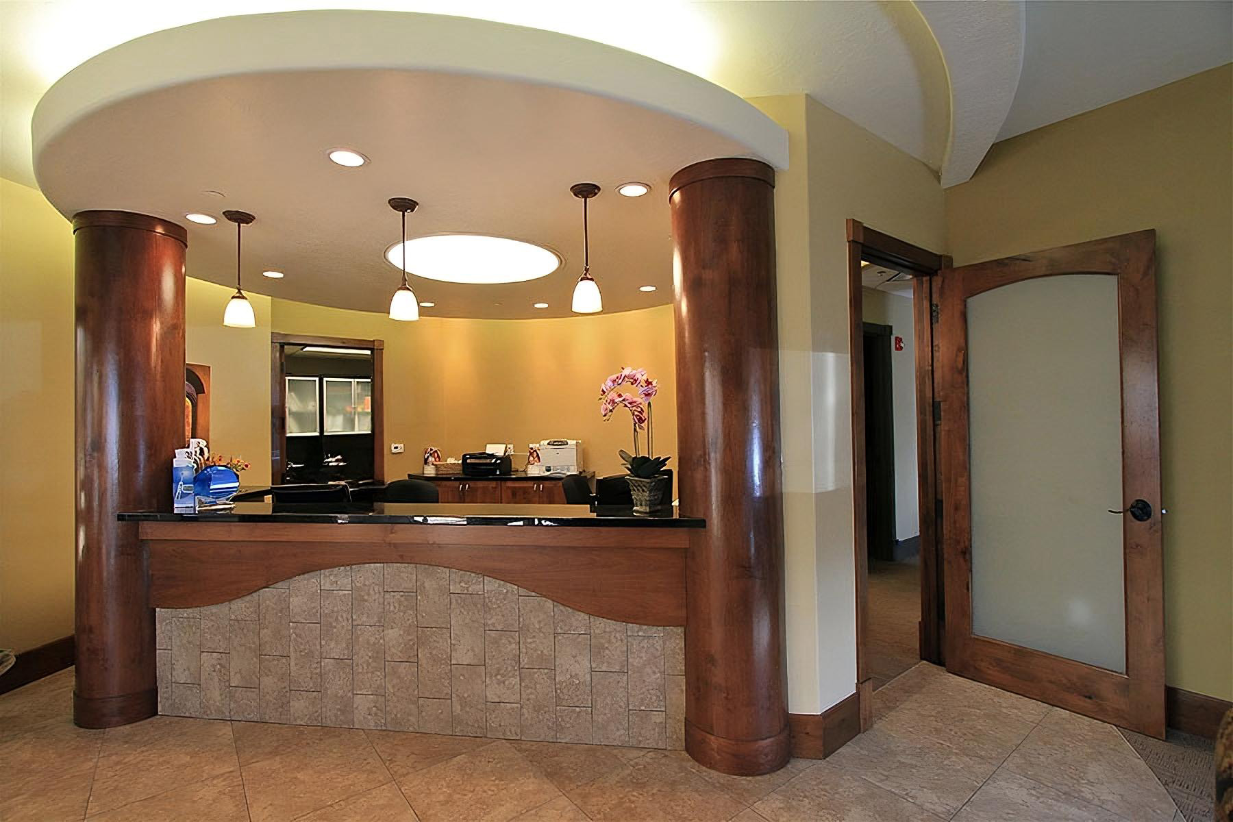 The image shows an interior view of a spacious lobby with a modern design, featuring a curved wooden countertop, a stone fireplace, a large window, and a beige wall with a decorative archway.