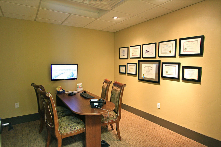 The image shows a room with a desk, chair, television screen displaying a presentation, and a wall adorned with framed certificates and awards.