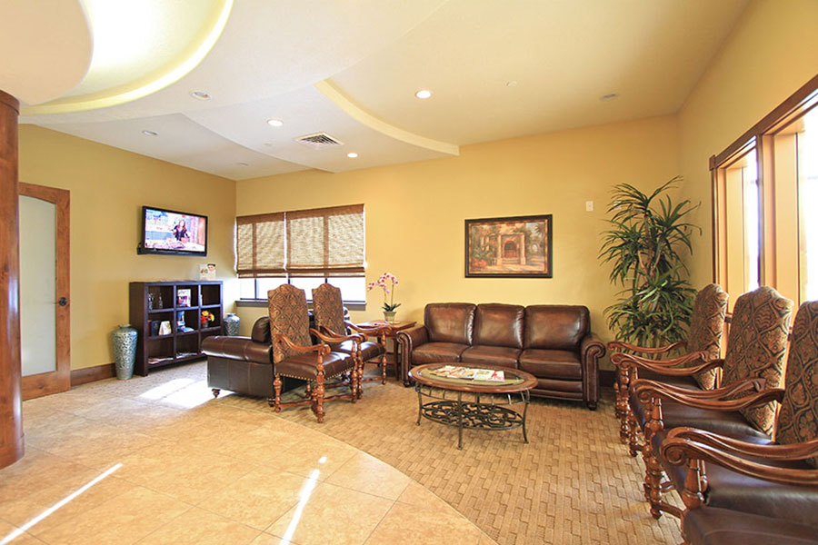 The image depicts an interior view of a well-lit waiting area within a building, featuring comfortable seating arrangements with leather upholstery, a large window allowing natural light, and a modern decor style with neutral colors and a framed picture on the wall.