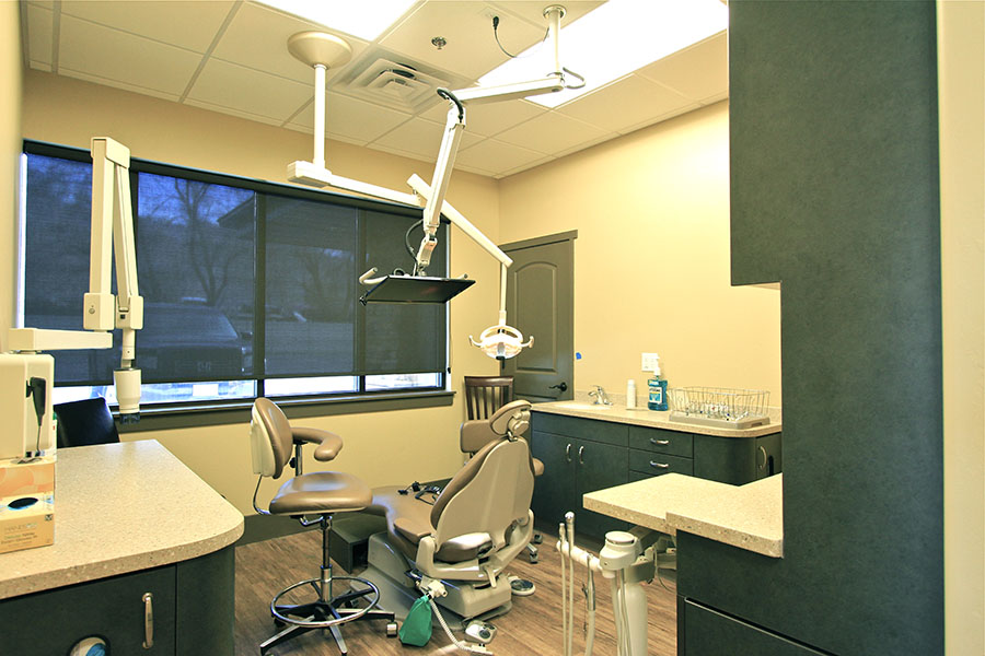 The image depicts a well-equipped dental office with a dentist's chair, examination equipment, and various dental tools visible.
