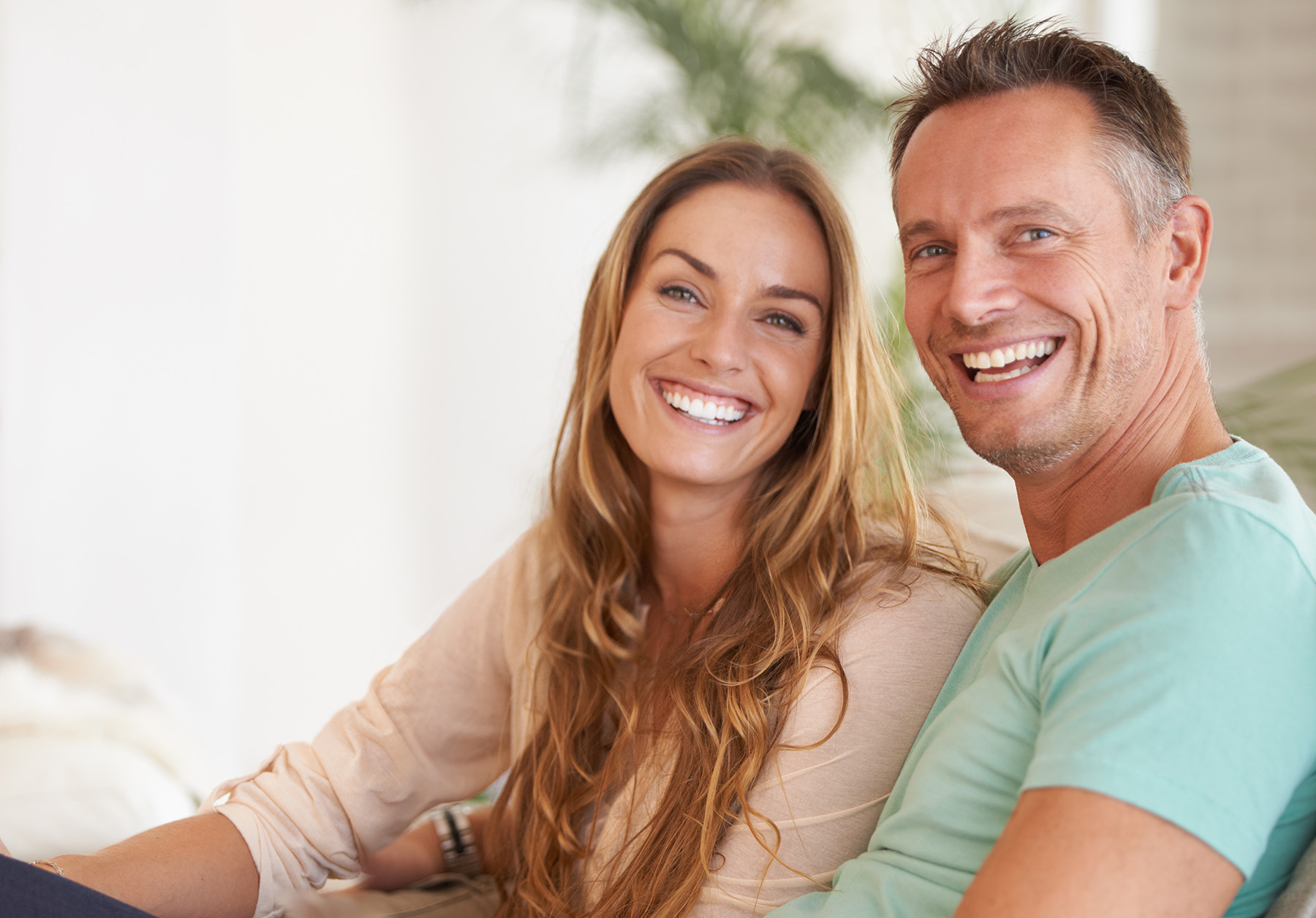 In the image, there are two adults, a man and a woman, sitting closely together on a couch, smiling at the camera, with the man wearing a blue shirt and the woman wearing a white top.