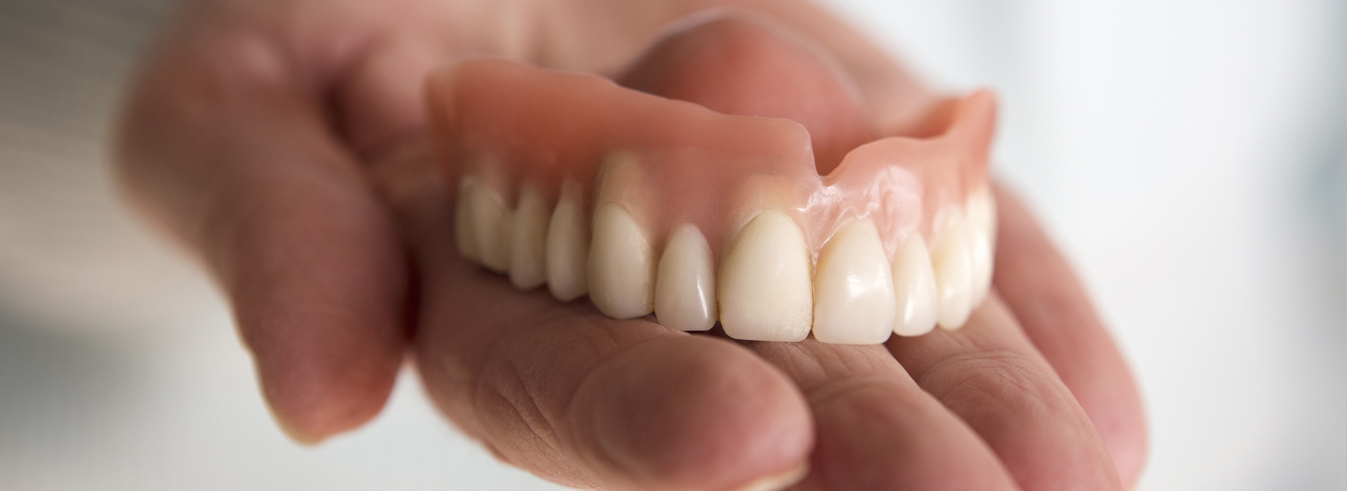 An elderly hand holding a set of dentures with open mouths, against a blurred background.