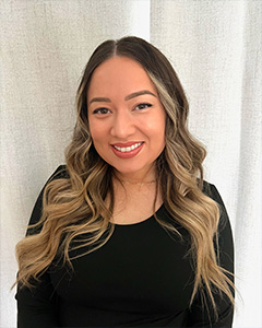 The image shows a woman posing with a smile, wearing makeup, long hair, and a black top. She has her hair styled down and is looking directly at the camera.