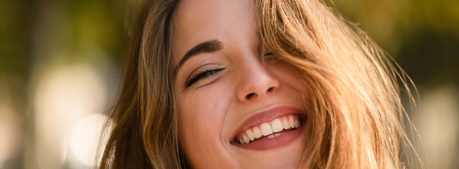 The image shows a woman smiling at the camera with her head slightly tilted, set against an out-of-focus background.