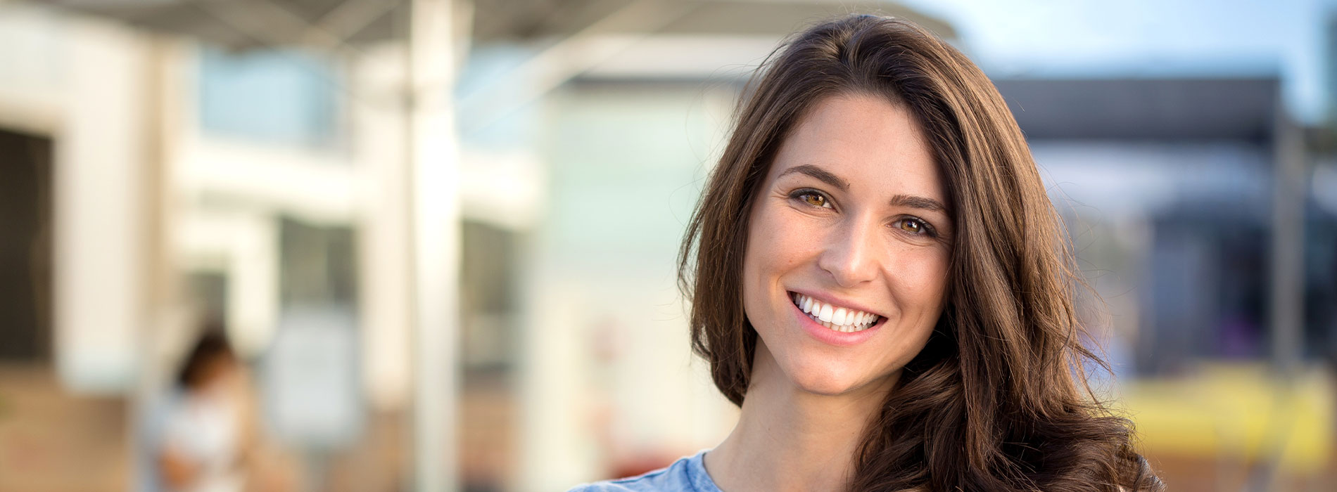 The image shows a smiling woman with long hair, wearing a blue top, standing outdoors during daylight.