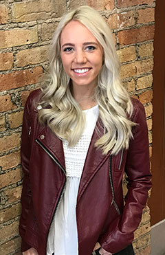 A woman with blonde hair wearing a red leather jacket and standing against a brick wall.