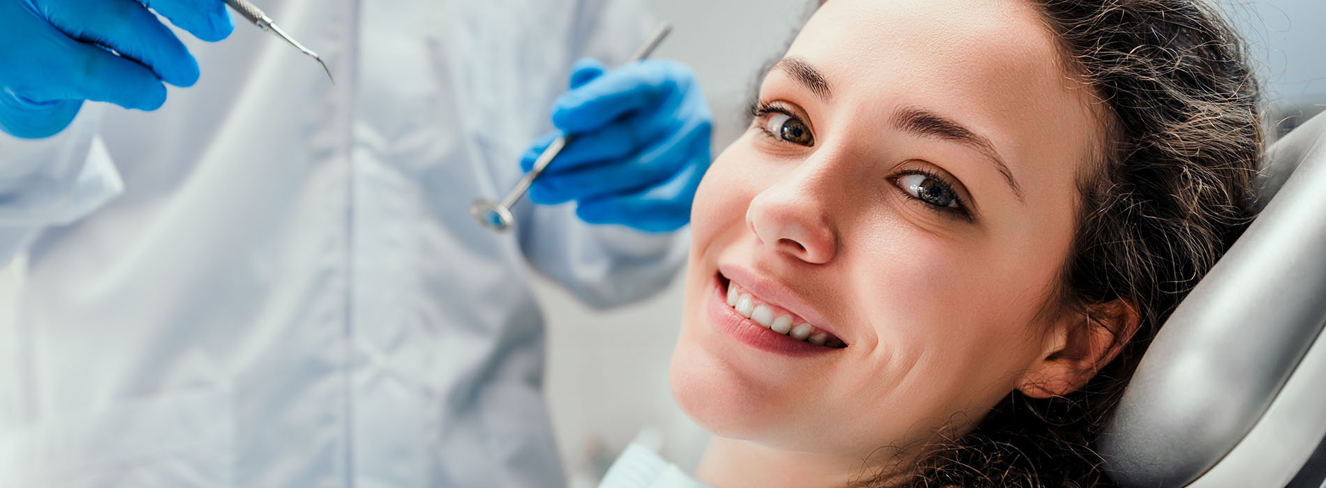 The image shows a woman smiling at the camera while seated in a dental chair with blue gloves on her hands, suggesting she is undergoing dental treatment.