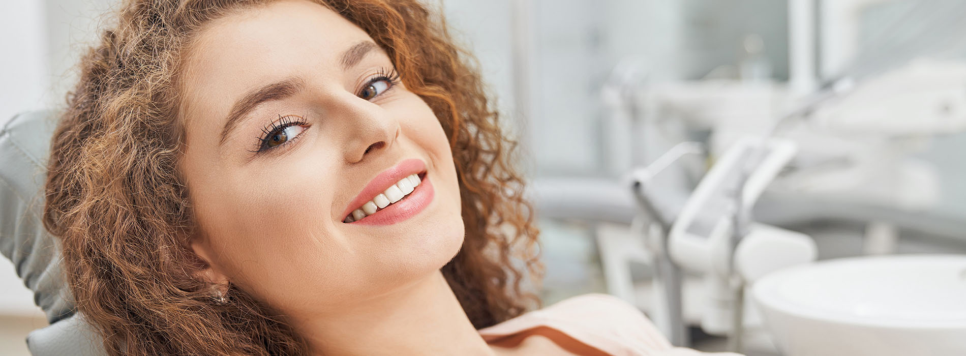 The image features a smiling woman sitting in a dental chair with her eyes closed, holding up a hand gestureing happiness or excitement, while a dental professional works behind her.