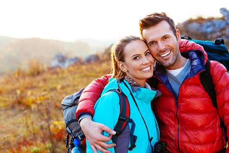 The image shows a man and woman embracing each other outdoors during daylight, with both wearing backpacks and outdoor attire suitable for hiking or adventure travel.