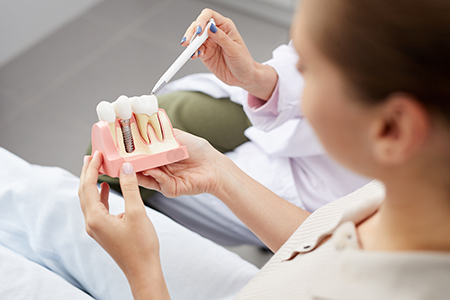 The image shows a person holding a model of a human mouth with teeth and gums, while another individual appears to be observing or assisting the first from a dental chair, suggesting an educational or demonstration setting related to dental care or education.
